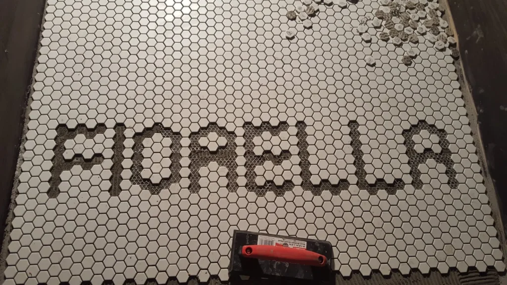 Close-up of the FIORELLA letter openings in white hexagon mosaic during installation, with removed tiles set aside.