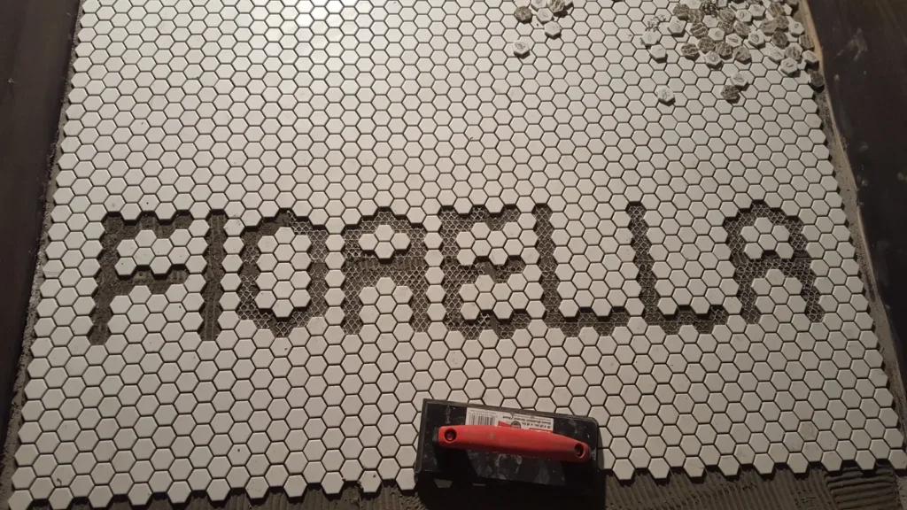 Letter-shaped openings for FIORELLA cut into white hexagon mosaic sheets before the dark lettering tiles are set.