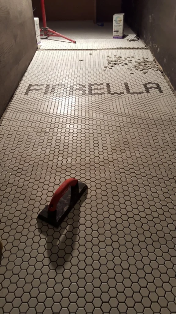 Long view of custom Fiorella lettering integrated into white hexagon mosaic restaurant flooring during installation.