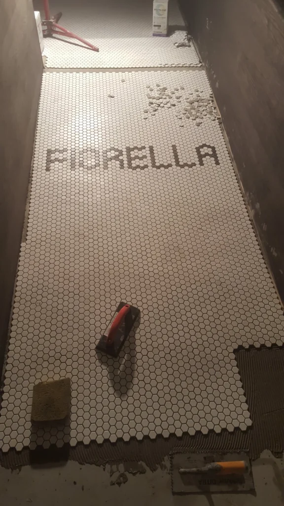Dark hexagon mosaic tiles spelling FIORELLA against a white tile floor during custom restaurant tile installation.