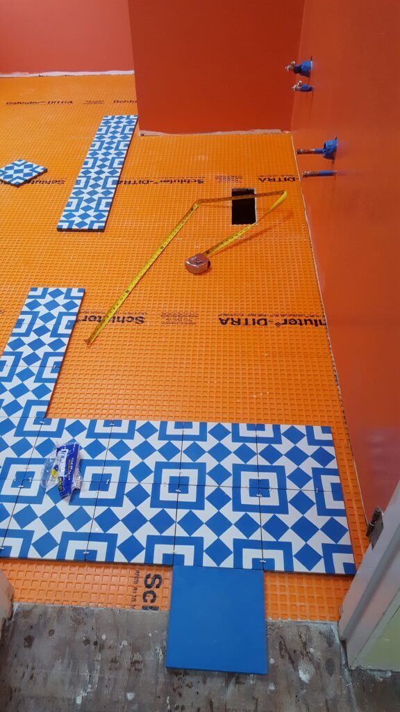 Dry layout of blue-and-white geometric floor tile on orange Schluter-DITRA membrane at the doorway of a Mill Valley bathroom, with a solid blue border tile and tape measure.